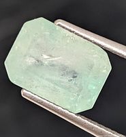 Genuine light green emerald gemstone with emerald cut shown in tweezers with a dark background, visible natural inclusions noted.