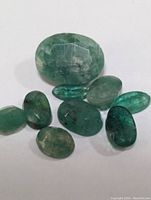 Set of eight loose emerald gemstones shown from overhead on white background, displaying color, shape, and natural inclusions.