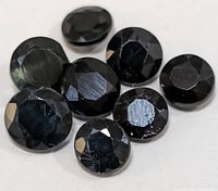 Close-up photo of eight faceted black sapphire gemstones on white background showing round cuts and reflective surfaces.