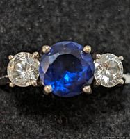 Close-up photo of the sterling silver ring showing the round deep blue faceted center gemstone flanked by two smaller round clear gemstones set in prongs.