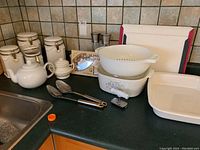 Photo showing collection of kitchen items including ceramic teapot, canisters, sugar bowl, colander, mixing bowl, salt and pepper shakers, cutting board, serving utensils, and metal condiment holder.