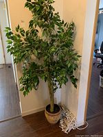 Front view of artificial tree in beige pot with foliage and a bundled string of white decorative lights on the floor next to it.