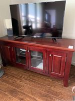 Front angle view of dark reddish-brown wooden cabinet with flat top, housing TV, lamp on sides, with two glass-paneled central doors and two solid wood side doors.