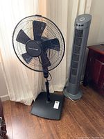 Photo of Bionaire black pedestal fan and gray tower fan side by side on wooden floor near cream curtains.