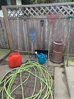 Full view of all gardening tools and accessories arranged against a wooden fence including rakes, a garden fork, a snow shovel, watering cans, garden hose, gas can and ceramic planters.