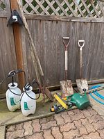Photo showing the full lot of garden tools arranged against a wooden fence, includes two shovels, two sprayers, hose nozzle, brush, hose and a hoe.