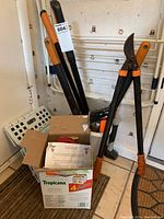 Gardening tools including long-handled Fiskars shears with orange handles, a black axe with orange handle, a white bucket partly containing lamp base and other tools, photo showing lot contents and condition.