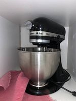 Front angled view of black KitchenAid Classic stand mixer with stainless steel mixing bowl on base with power cord and red cloth nearby.