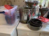 Photo showing large stainless steel pots, round baking pans, metal strainer with wooden pestle, and storage bin with plastic Tupperware containers.