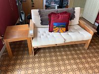 Photo showing wooden frame loveseat with cream tufted cushions and two red pillows, gray throw blanket draped over backrest. Wooden side table next to loveseat.