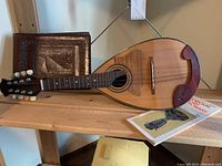 Mandolin laid horizontally with a leather bound photo album upright behind it and a mandolin instruction booklet to the right on a wooden shelf.