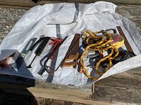 Photo of assorted outdoor tools on white surface including jumper cables, hand saws, crowbar, and yellow orange rope.