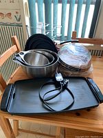Overview of the electric grill, nested stainless steel mixing bowls, stacked frying pans, plastic-wrapped lids, and metal lid rack on a wooden table.