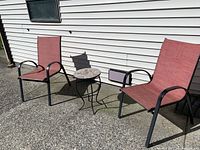 Two metal-frame patio chairs with red fabric seats and a small round metal table with mosaic tile top arranged outside against white siding.