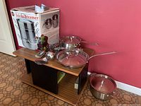 Photo shows stainless steel pots and pans on shelf including a saucepan with lid, frying pan/skillet with glass lid, small pot with lid, and boxed juicer/steamer unit with various kitchen utensils.