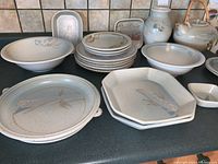Full view of pottery collection showing stacked plates, bowls, pitchers, and teapots with bamboo handles in soft blue-green decor.