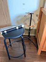 Wooden tambourine on black guitar stand next to black round seat stool with tripod flute on stool seat
