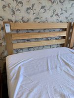 Twin wooden bed frame with horizontally slatted headboard in light natural wood finish, portion of mattress covered by white fitted sheet visible.