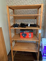 Front view of wooden shelving unit with four shelves. Shows box and camera tripod on shelves, wooden texture, and background floral wallpaper.