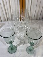 Photo of the 8 wine goblets, including 2 green and 6 clear, arranged together on a white surface with curtain background.