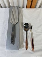 Photo showing three serving utensils on a white cloth background: a large Community server on a gray box, a smaller spoon and fork with copper-toned handles.
