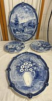 Four handpainted blue and white ceramic items displayed on fabric: two large oval platters with scenic and floral designs, two smaller plates with village scenes.