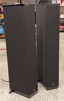 Pair of tall black fabric-covered Definitive Technology powered speakers standing side by side on a speckled floor with visible cables.