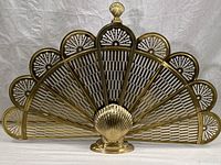 Front view of a brass folding fireplace screen, showing its fan shape with seven perforated panels and shell motifs on top of each.