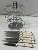 Photo showing set of five steak knives with cream-colored handles and one paring knife in front of a chrome rotating spice rack with 10 round slots