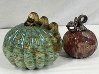 Two glass pumpkins, larger greenish with brown swirled pattern and curled stem, smaller reddish with curled stem.