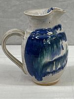 Front angle showing the glazed white ceramic body with prominent blue and green drip glaze pattern, large handle and pouring spout.