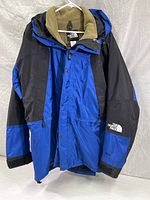 Full view of the blue and black The North Face winter jacket hanging on a white hanger, showing front zipper and snap button cover, with removable fleece lining partially visible inside.