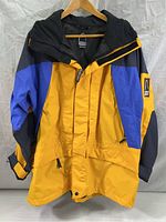 Full front view of the men's yellow, blue, and black Sierra Designs rain jacket with hood, hanging on a wooden hanger against a white backdrop.