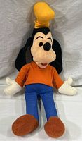 Front full view of large vintage Goofy plush toy showing orange shirt, blue pants, brown shoes and yellow hat