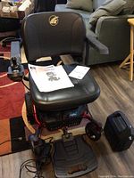 Front view of the electric wheelchair showing cushioned seat, joystick on right armrest, footrest, and one of two additional batteries beside it.