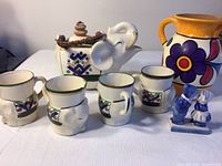 Full set of elephant tea set: white ceramic teapot shaped like an elephant with brown woven handle and four matching elephant-shaped cups with colorful geometric patterns, one cup has a small chip, small blue and white Dutch figurines labeled Holland, bright orange and purple flowered pottery pitcher from Cabo Mexico.