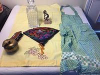 Entire lot displayed on yellow cloth including vintage apron, glass decanter, brass apple paperweight, opera glasses, Kokopelli on stone, and Japanese fan