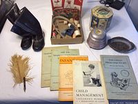 Photo showing a collection of old pamphlets, black child's rain boots, a box with sewing supplies, an ink well with feather pen, a small music box, and a decorative tray.