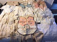 Overview of multiple ecru and white vintage baby clothes, blankets, embroidered bib, pillow case, and doilies laid out on a surface.