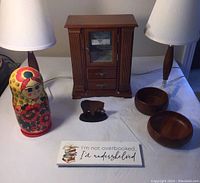 Photo of lot showing two small 1960s style lamps, wooden jewelry box with drawers and mirror, Russian nesting doll with floral design, two wooden bowls, small grizzly bear figurine holding fish, and desk sign with quote.