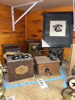 Five antique wooden box telephones arranged together with one framed art print showing a rotary phone image, placed on a wood shelf.