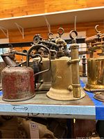 Four metal and brass blow torches arranged on a shelf, showing a variety of designs and conditions.