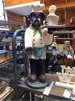Full frontal view of large dog chef figurine holding tray, showing details of outfit and face, standing on circular base.