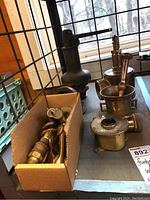 Wide shot showing multiple items on table including fire hose valve with lever, brass mortar and pestles, a cardboard box with brass valve parts.