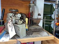 Small metal lathe and grinding wheel on wooden base next to a glass jar with candles and spools on shelf.