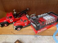 Three metal model trucks on wooden shelf: boxed True Value 1918 Mack flatbed truck, red Mobiloil tow truck, and red Texaco tow truck