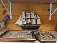 Photo showing wooden model ship Pamir centered with detailed sails and rigging, brass porthole on the right, and framed knot art on the left.