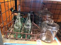Metal carrier filled with two vintage embossed Coca-Cola bottles and decorative figurines inside. Two clear glass lidded jars near the carrier.