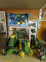 Six green and yellow metal toy tractors and a small metal forklift on wood shelf, with boxed models and wall sign in background