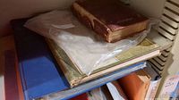 Stack of antique books including a large brown leather-bound book on top, a plastic sleeve with prints underneath, another green book, and a blue binder at the bottom.